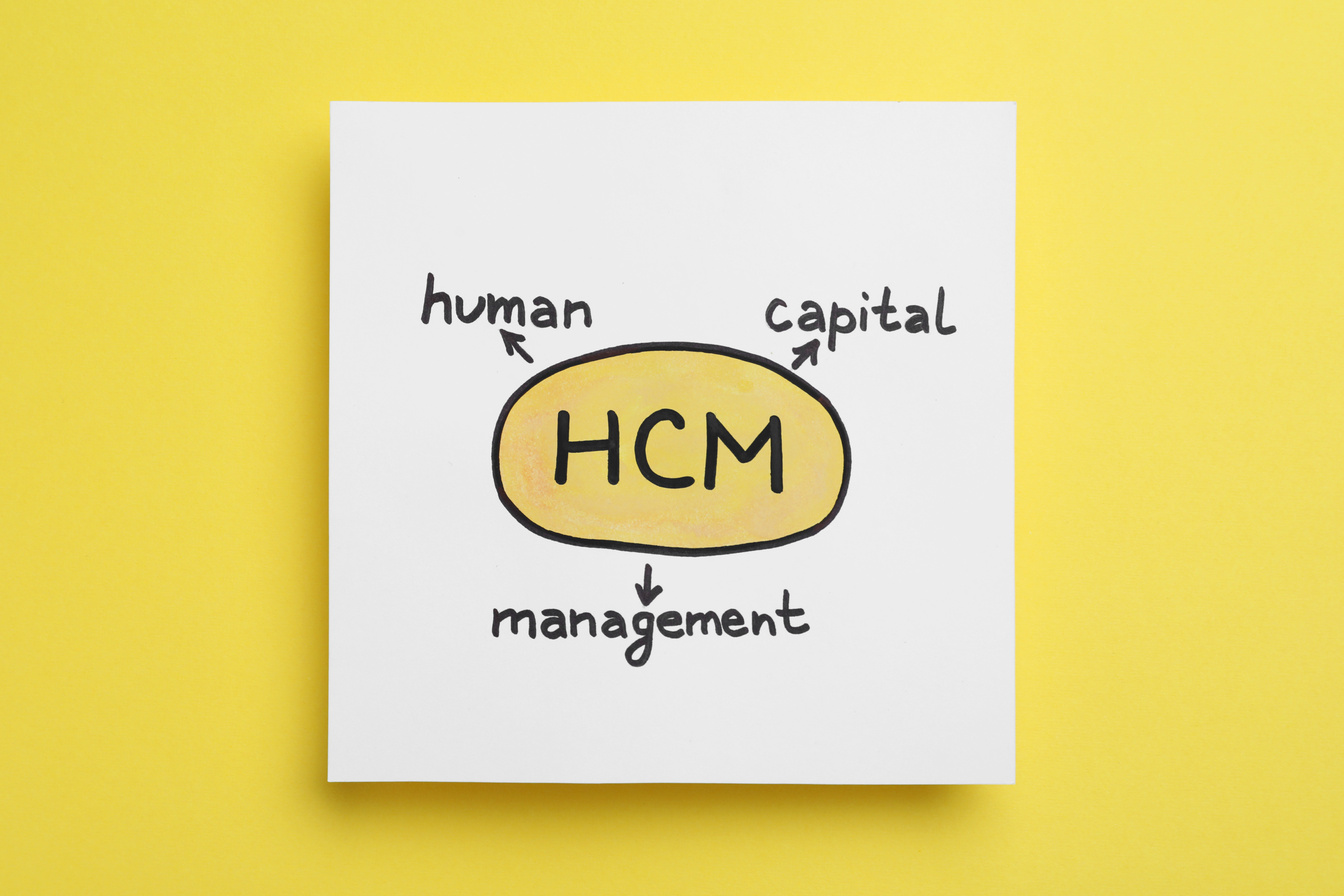 Paper with HCM Abbreviation and Its Interpretation (Human Capital Management) on Yellow Background, Top View
